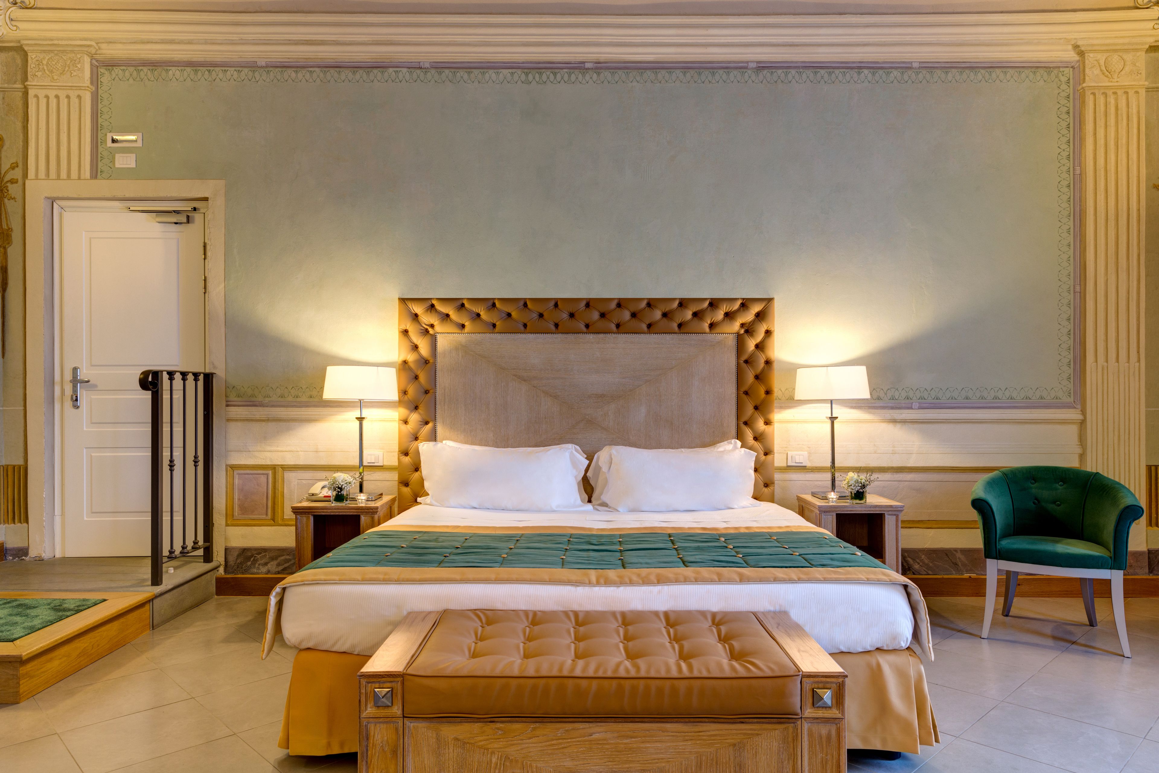A large bed with a tufted headboard, two bedside lamps, a green armchair, and a wooden bench.