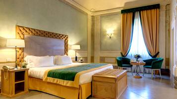 Habitación de hotel con una cama grande, dos lámparas de noche, una mesita de noche de madera, una pequeña mesa con un florero y dos sillones verdes.