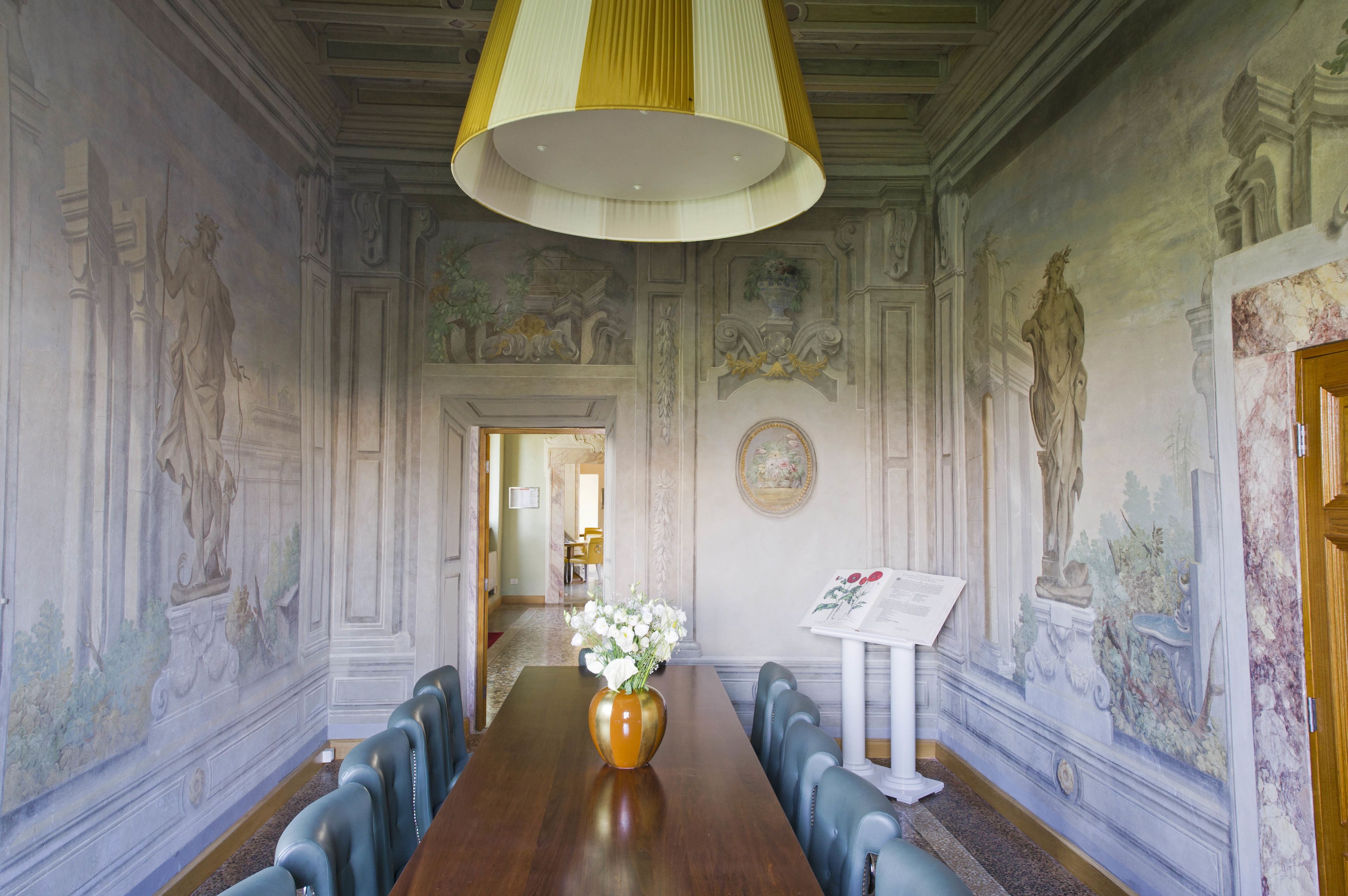 A formal dining room with a long table, blue upholstered chairs, a large vase with flowers, and classical wall murals.