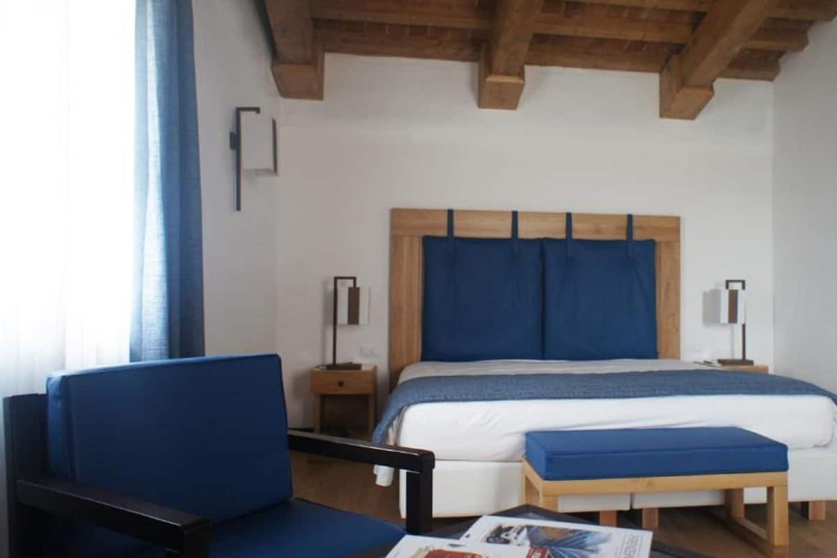 A bedroom with a wooden headboard, a bed with blue bedding, a bedside table with a lamp, a chair, and a wooden bench.