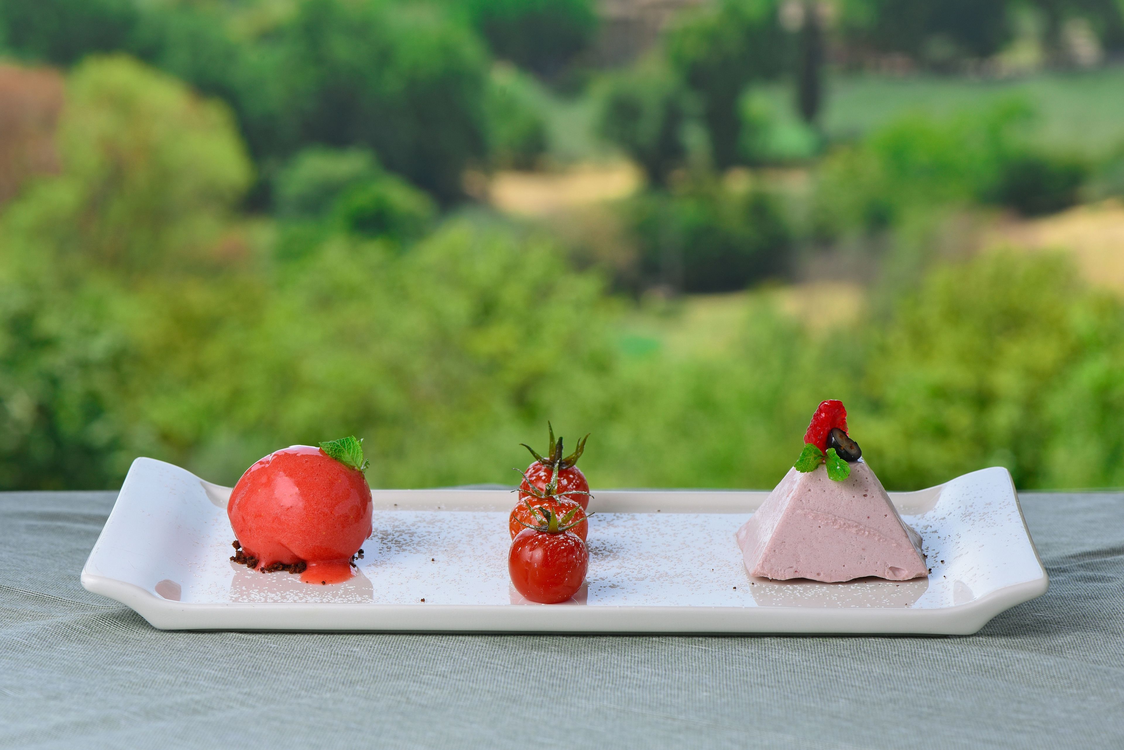 Ein Dessertteller mit drei Elementen: einer roten Kugel, einer kleinen Tomate und einem rosa Eisbecher, vor unscharfem Grün.