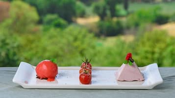 Ein Dessertteller mit drei Elementen: einer roten Kugel, einer kleinen Tomate und einem rosa Eisbecher, vor unscharfem Grün.