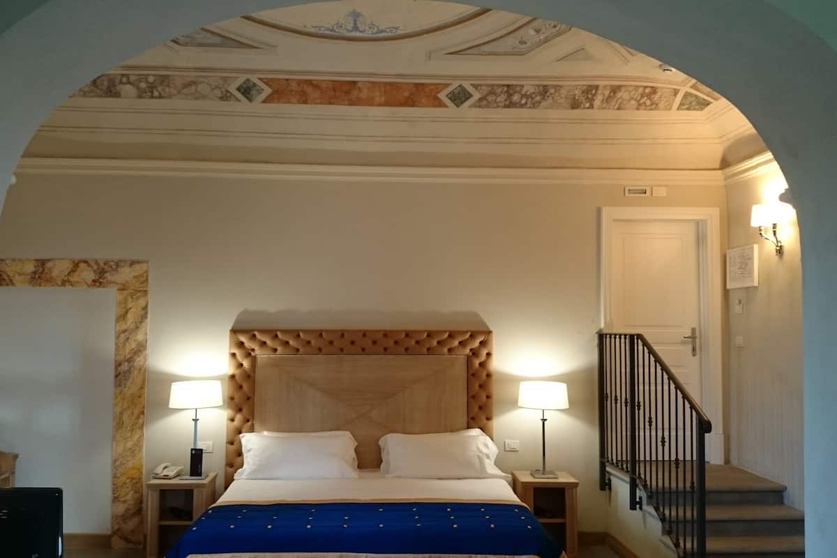 A hotel room with a bed, two bedside lamps, a wooden headboard, a marble wall, a staircase with a black railing, and a door.