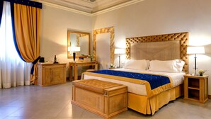 Une chambre spacieuse avec un grand lit, une tête de lit en bois et un pouf assorti. On y trouve une table de chevet en bois avec une lampe, une commode et un miroir. La chambre présente un style classique, avec un sol carrelé et un plafond décoratif.