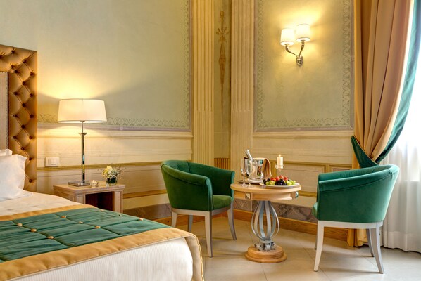 A hotel room with a large bed, two green armchairs, a small table with a fruit bowl, a lamp, and a mirror. - Villa Tolomei Hotel & Resort (Florence)