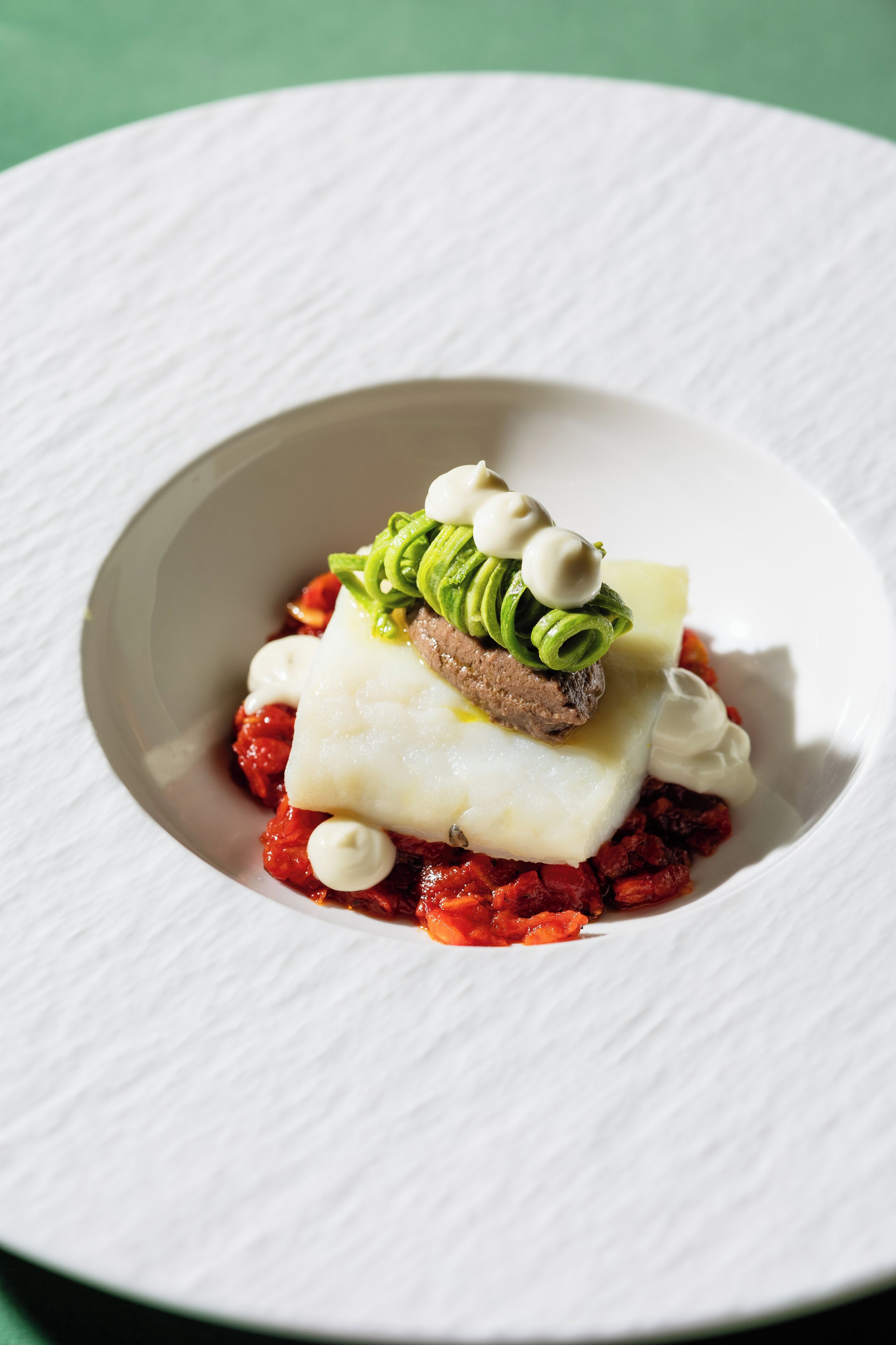 A white plate with a dish consisting of a white fish fillet, green avocado slices, and a piece of bread, served on a bed of red sauce.