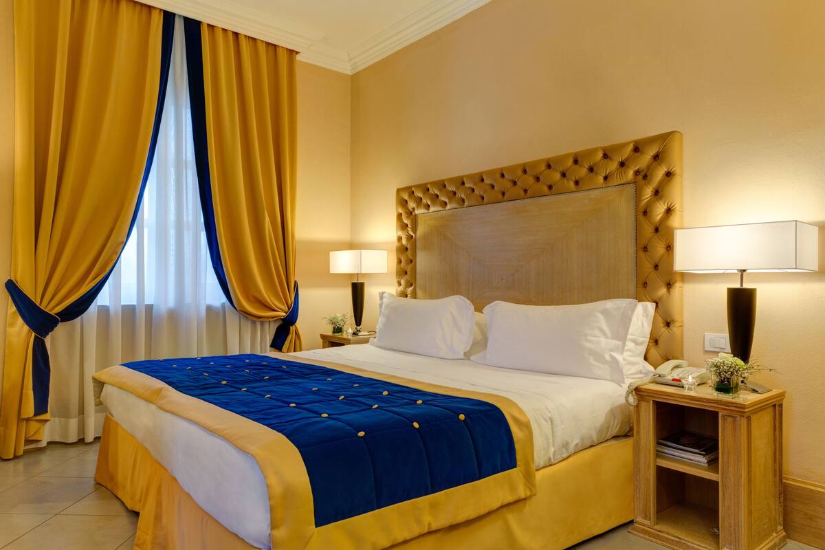 A hotel room with a large bed, two bedside lamps, a nightstand with a lamp, and a window with curtains.