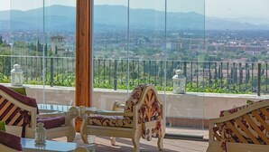 Un balcon avec un lit et des chaises en bois, d’où l’on peut admirer un paysage urbain avec des montagnes à l’horizon.