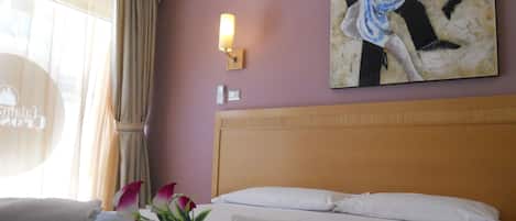 A hotel room with a bed, a wall-mounted air conditioner, a painting, a wall-mounted light, and a window with curtains.