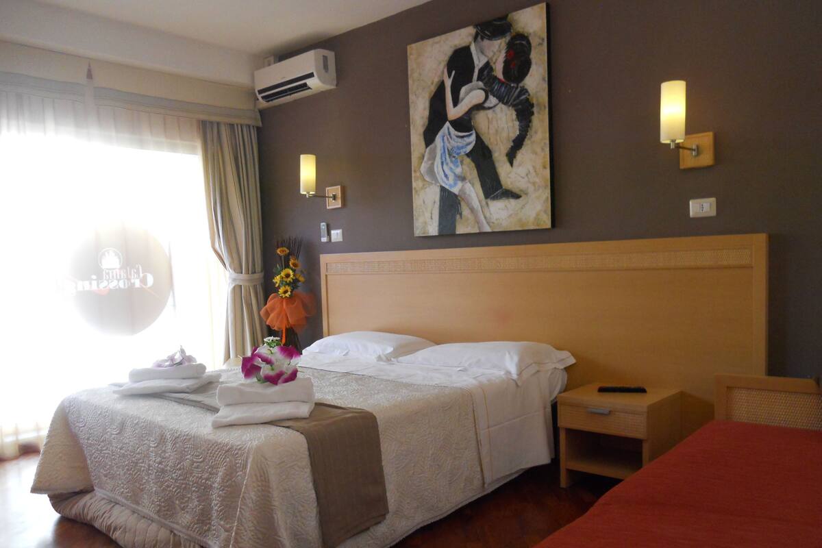 A hotel room with a large bed, a bedside table, a nightstand, an air conditioning unit, a painting on the wall, and a window with curtains.