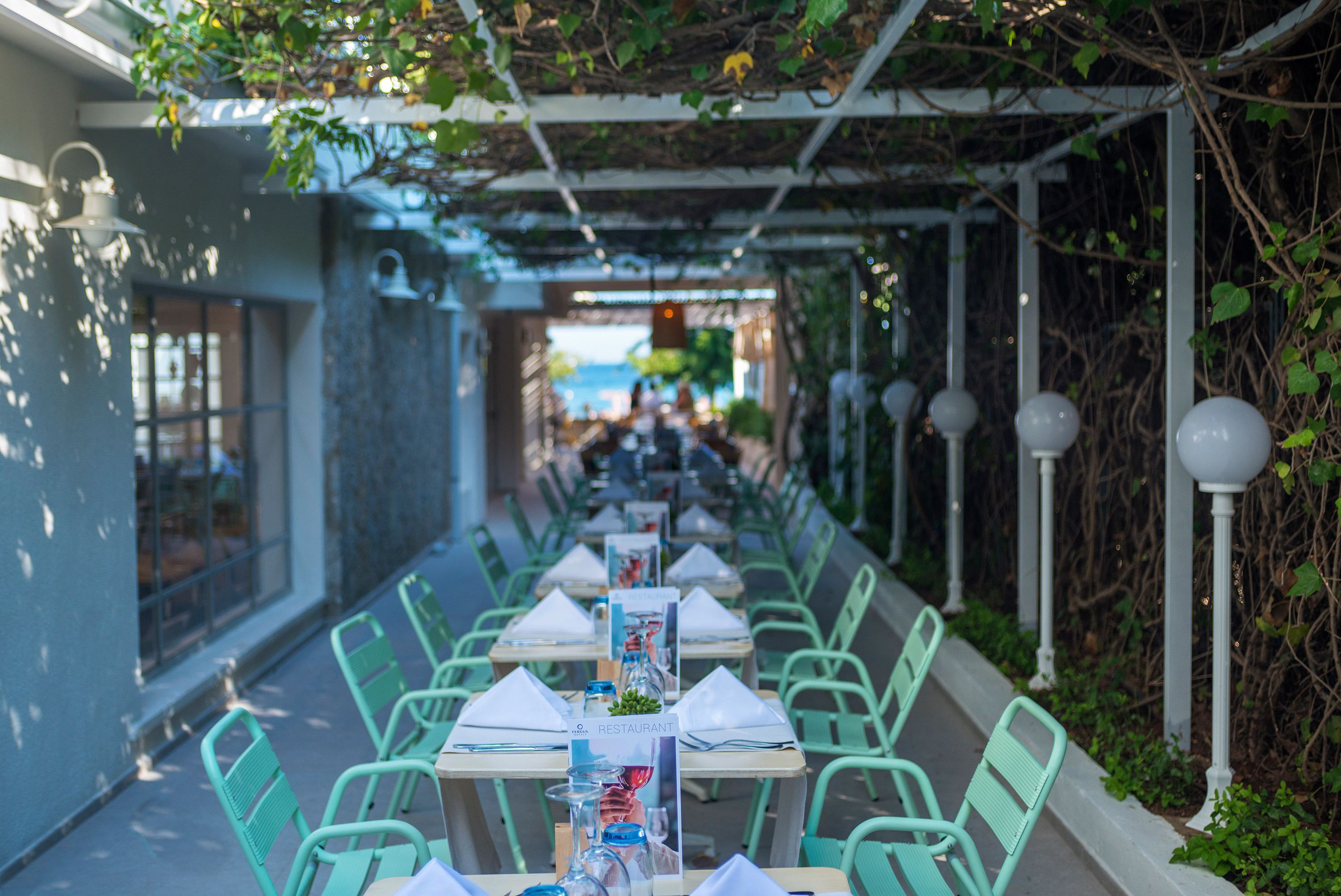 A covered outdoor dining area with tables and chairs, a pergola overhead, and a view of the street in the background.