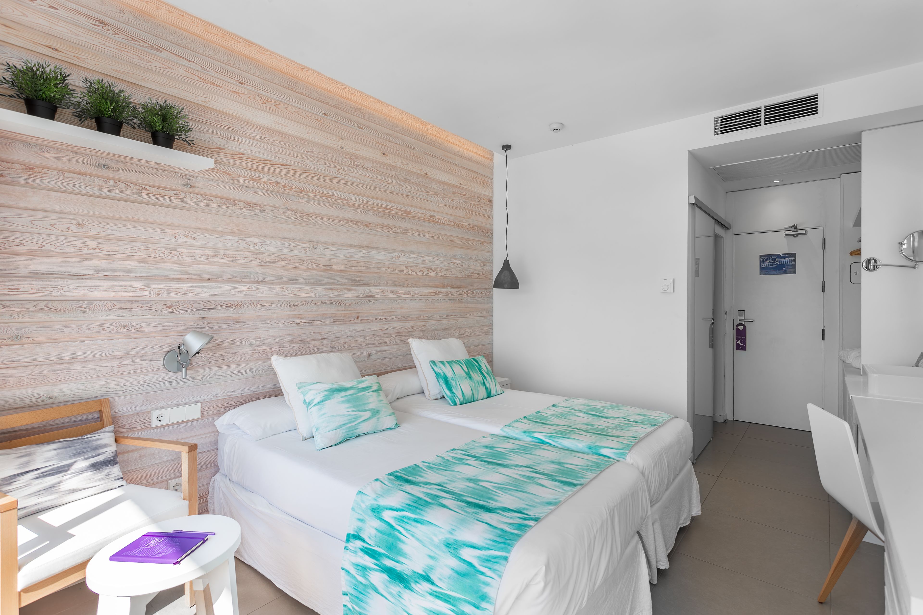 Un dormitorio moderno con una pared de acento de madera, una cama con colcha verde azulada y blanca, un escritorio con silla y una pequeña mesa con un cuaderno morado.
