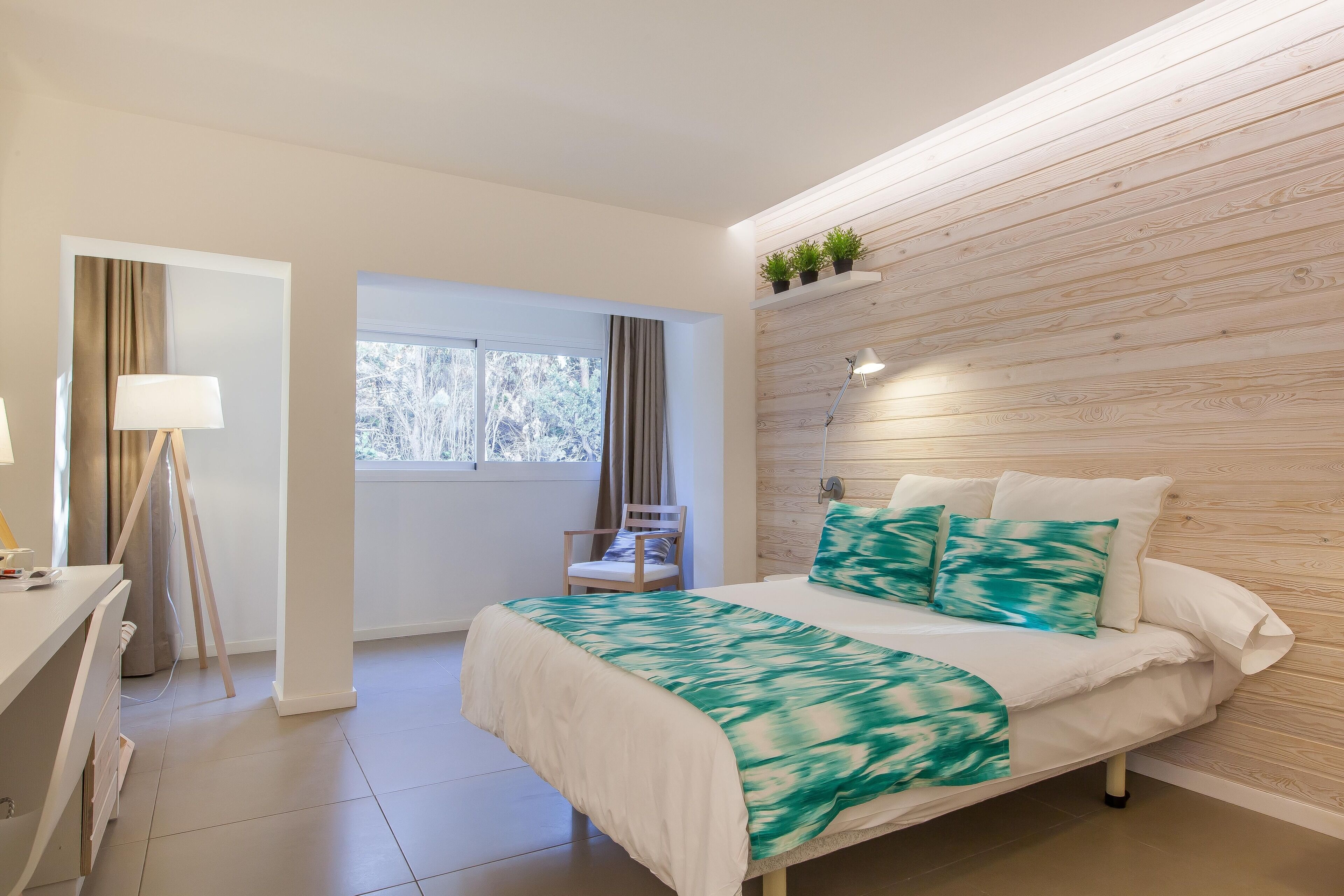 A bedroom with a bed, a desk, a chair, a window, and a wall-mounted shelf with plants.