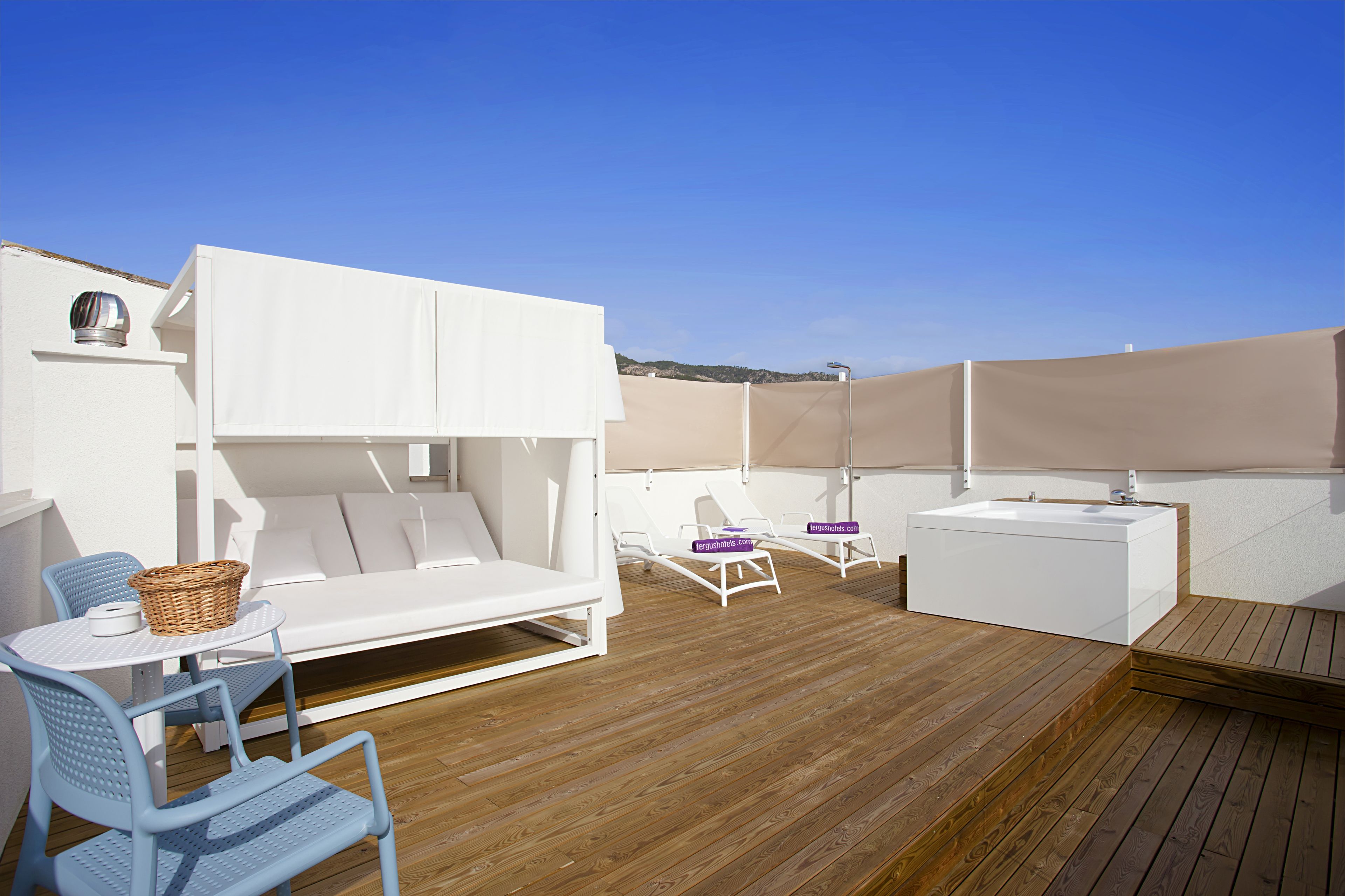 Une terrasse extérieure baignée de soleil, avec un canapé blanc, deux fauteuils de relaxation, une petite table surmontée d’un panier et une plateforme en bois.