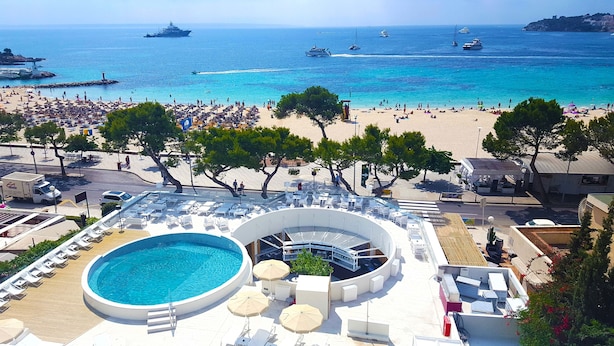 A poolside area with lounge chairs and umbrellas, overlooking a beach with people and boats. - FERGUS Style Palmanova - Adults Only (Calvia)