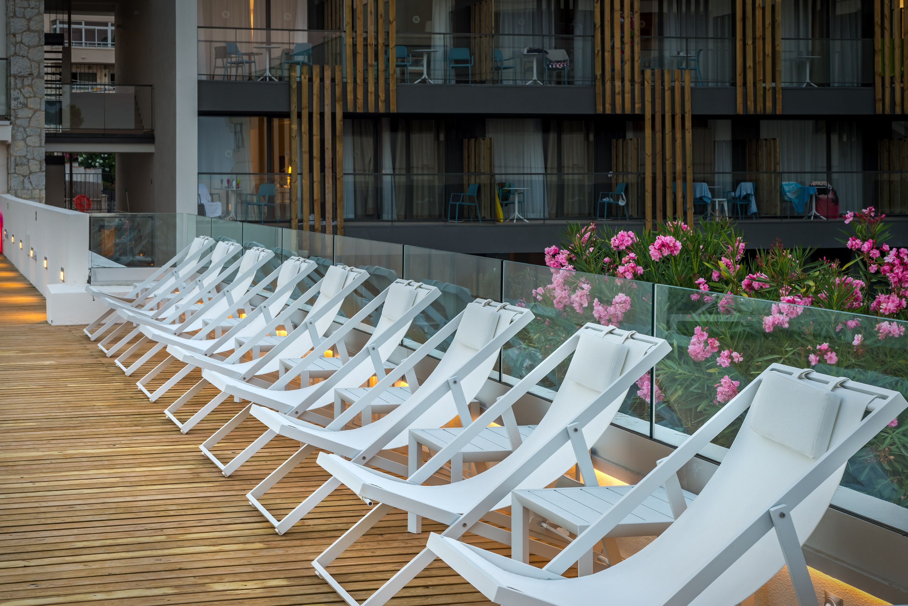 Une terrasse dotée de chaises longues blanches et d’une table en verre, offrant une vue sur un immeuble avec de nombreux balcons et des espaces de détente en plein air.