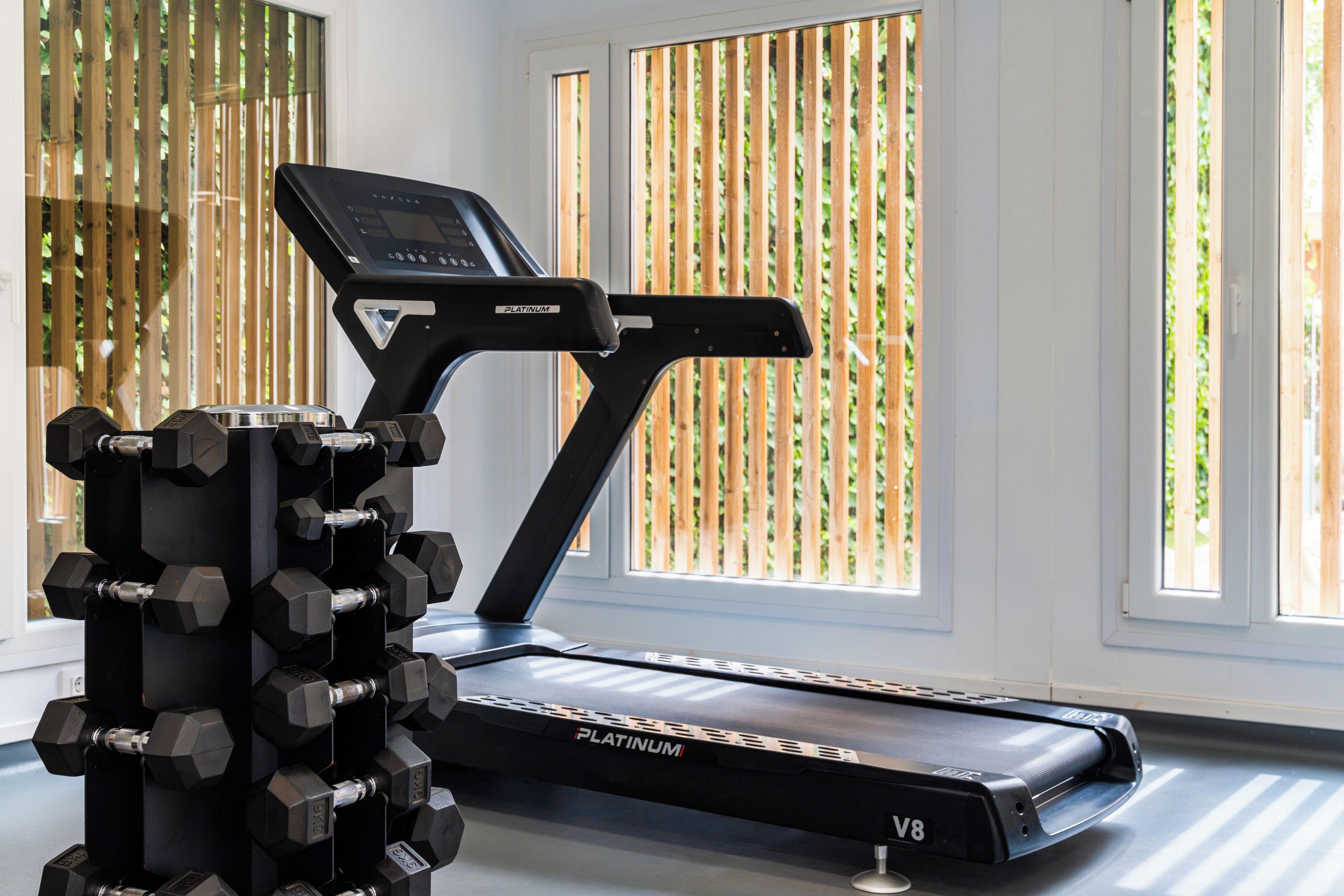 A treadmill with a digital display and a set of adjustable dumbbells in a room with large windows and a bamboo fence outside.