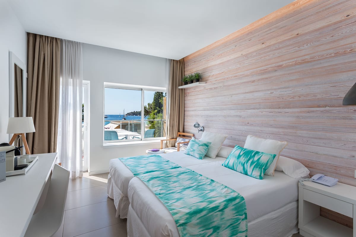 A modern hotel room with a large bed, a desk with a lamp, and a view of the sea through the window.