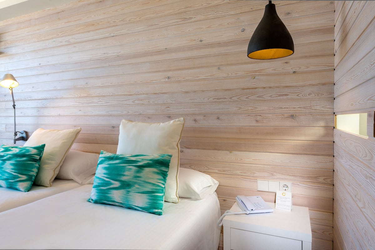 A bedroom with a wooden wall, a bed with white bedding and green pillows, a bedside lamp, and a nightstand with a phone and a sign.