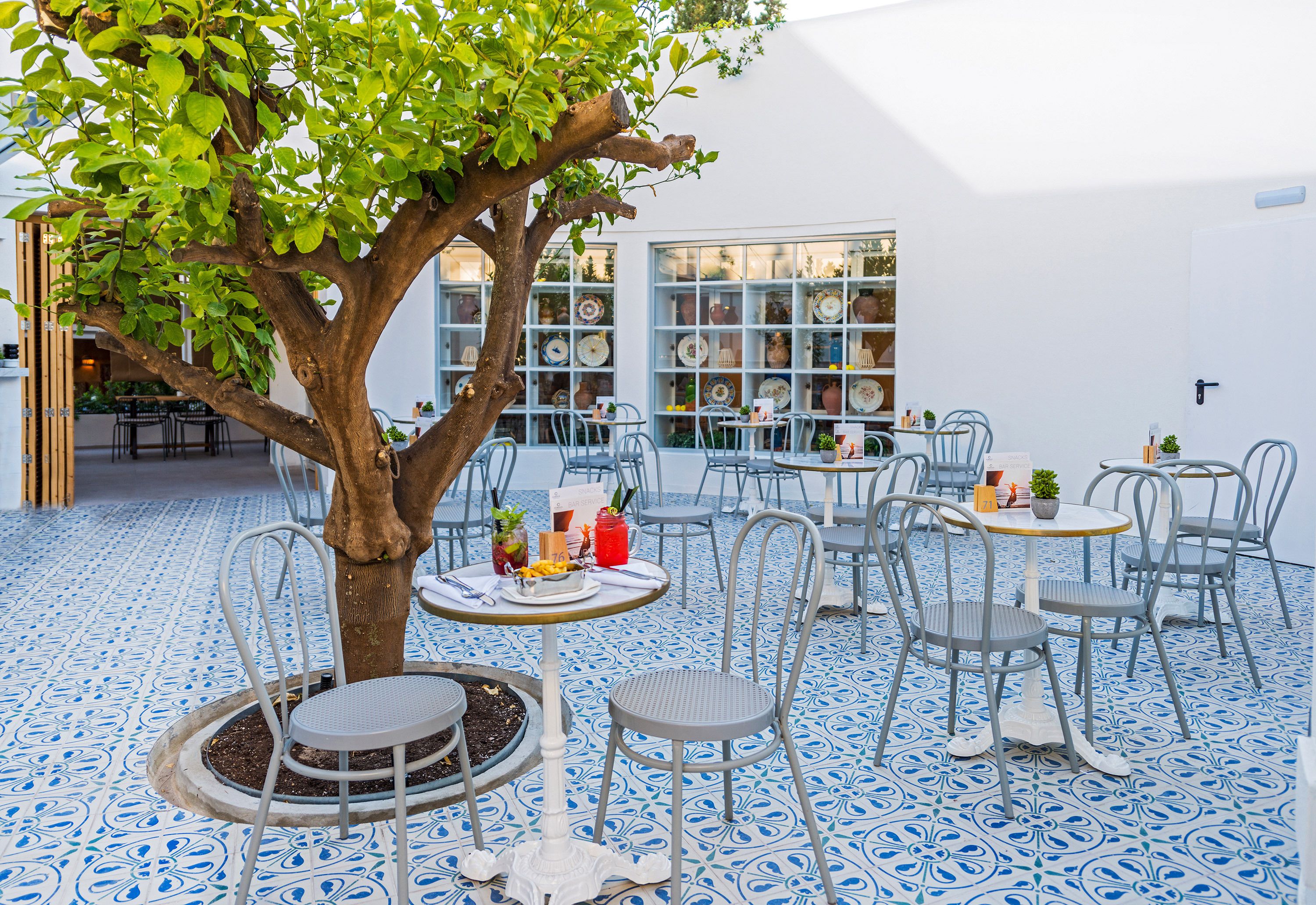 An outdoor café with tables and chairs, a tree, and a wall displaying various items.