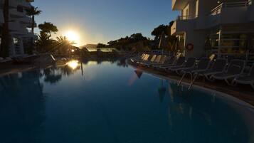 Uma área à beira da piscina com espreguiçadeiras e guarda-sóis, refletindo o pôr do sol.