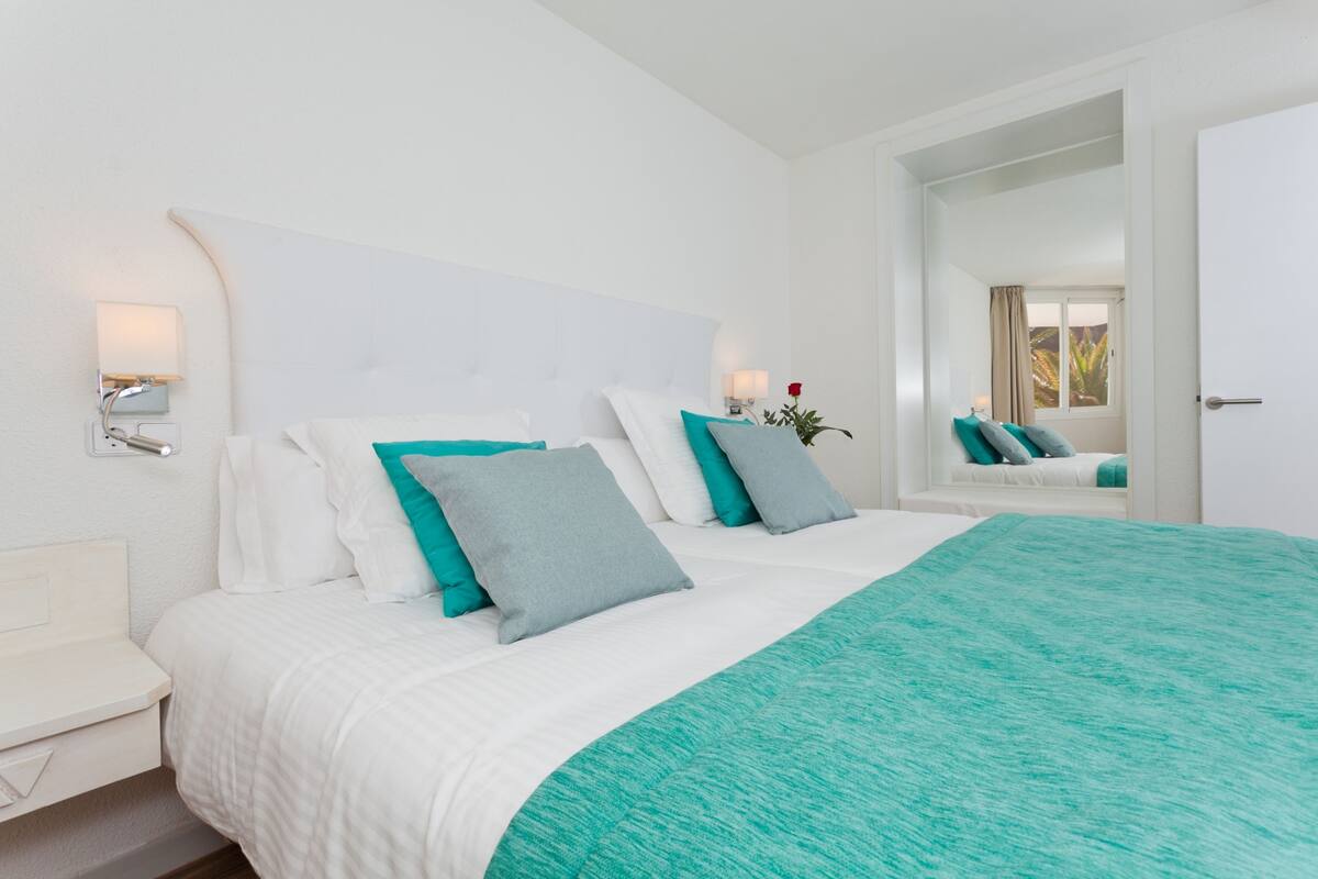 A neatly arranged bedroom with a large bed, white walls, and a mirror reflecting the room.