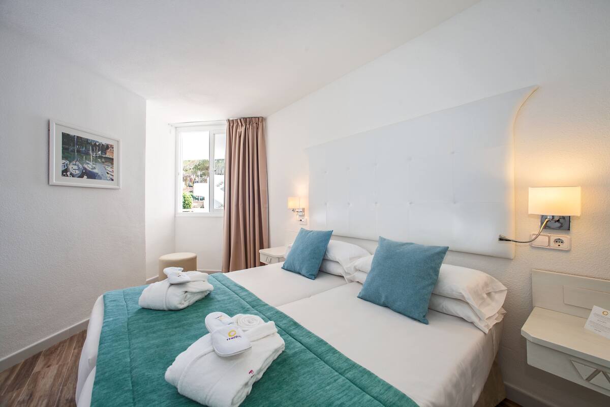 A hotel room with two beds, a window with curtains, a bedside table with a lamp, and a framed picture on the wall.