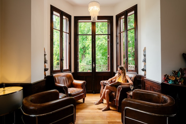 A room with a large bay window, leather armchairs, a wooden floor, and a chandelier.