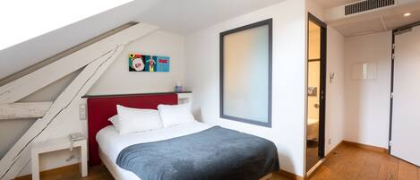 Un dormitorio con cama, cabecera roja, mesita de noche blanca, suelo de madera, tragaluz y un cuadro en la pared.