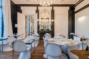 A formal dining room with white chairs and tables, a chandelier, and a fireplace.