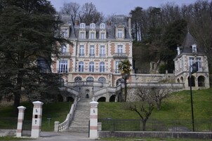A grand, multi-story building with a central staircase leading to an entrance, surrounded by lush greenery and trees.