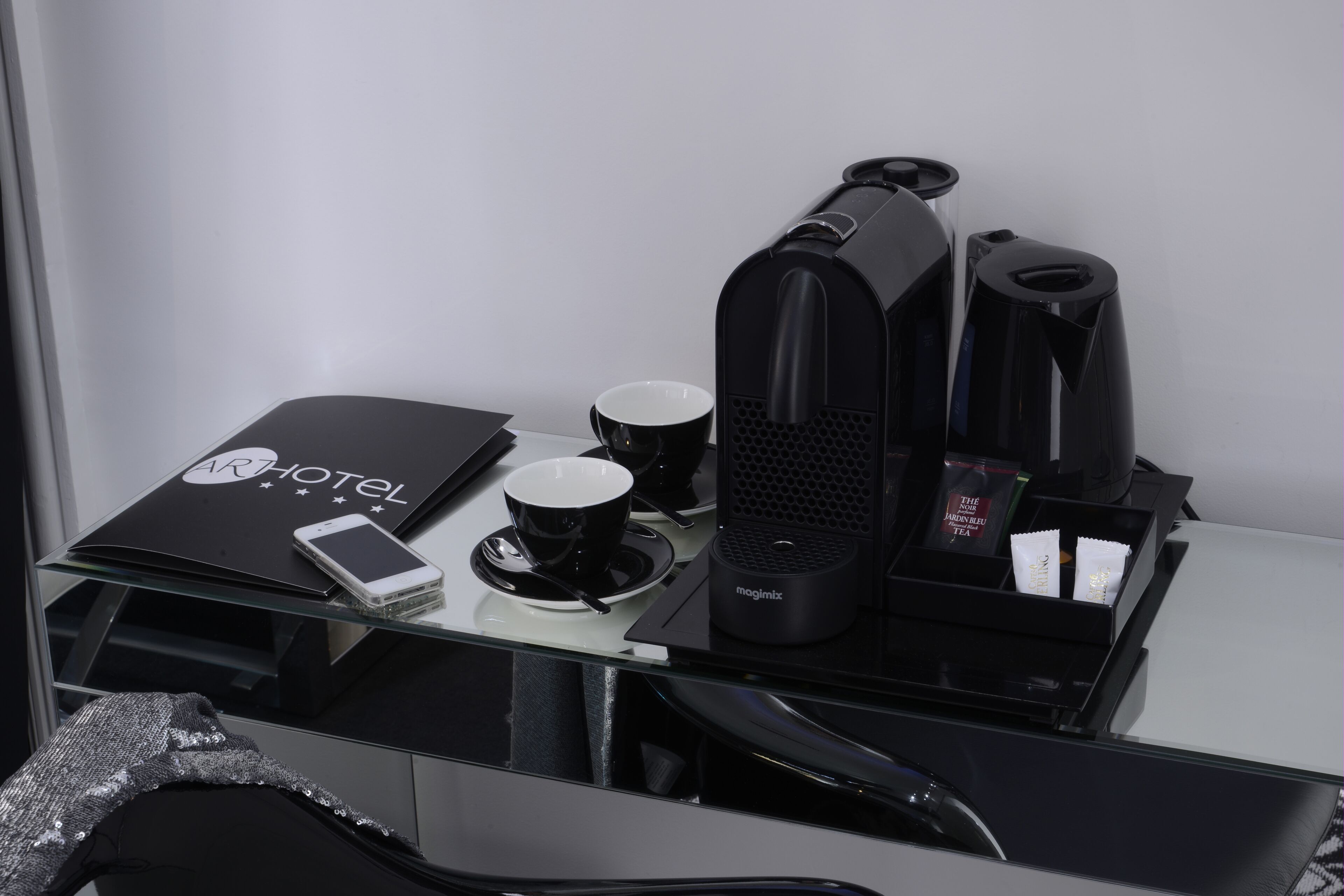 A hotel room with a glass desk, a smartphone, a mug, a coffee maker, and a tea set.