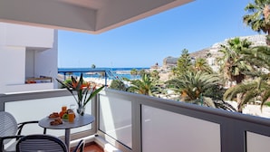A balcony with a table set for breakfast, overlooking a beach and a mountainous area. - Apartamentos Miami (Mogan)