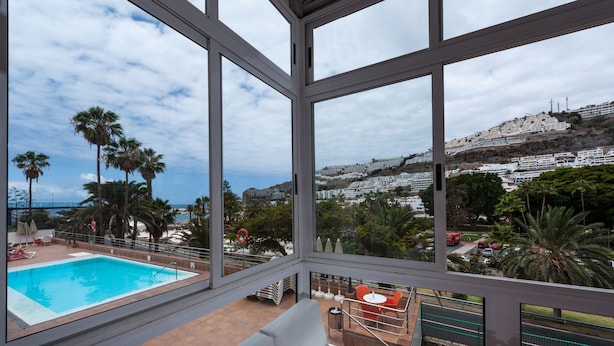A pool area with a view of a coastal town, including a beach and buildings on a hillside. - Apartamentos Miami (Mogan)