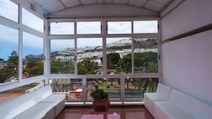 A sunlit room with large windows offering a view of a coastal town and greenery. - Apartamentos Miami (Mogan)