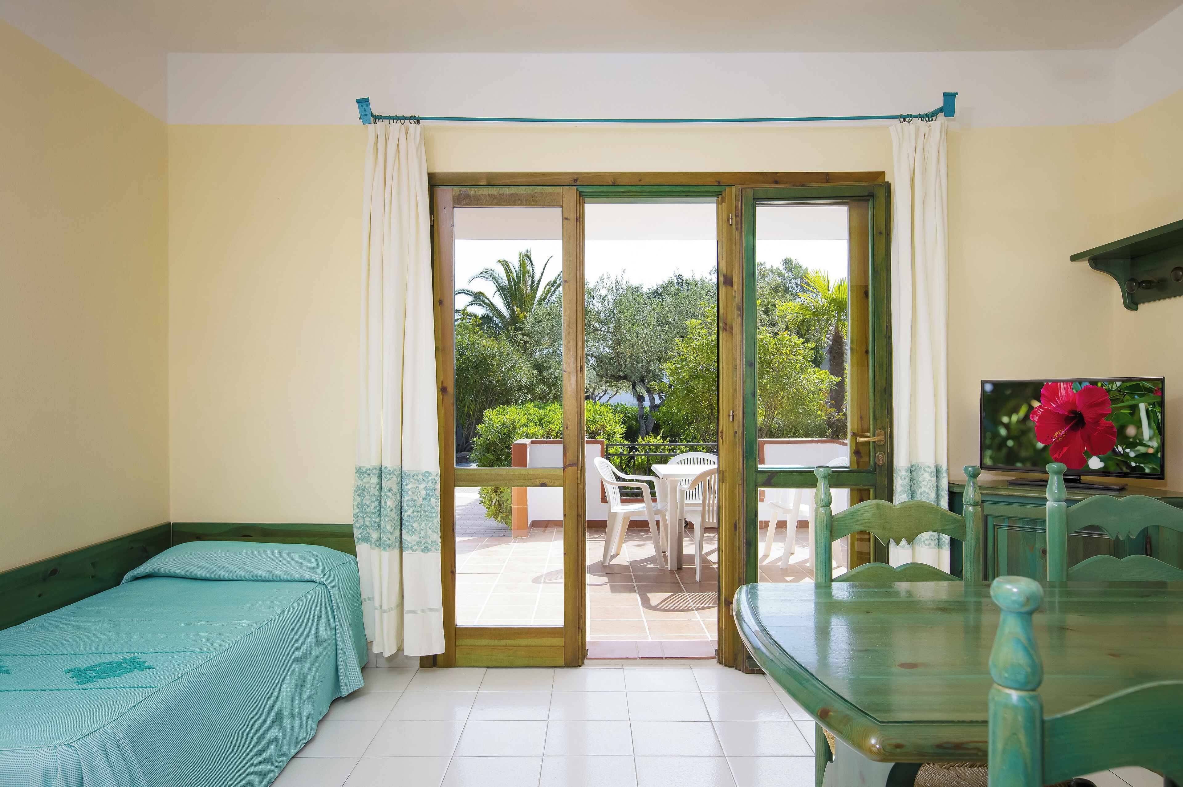 Una habitación con cama, mesa y silla, puerta corrediza de vidrio que da a un espacio exterior con mesa y sillas, y un televisor sobre un mueble.