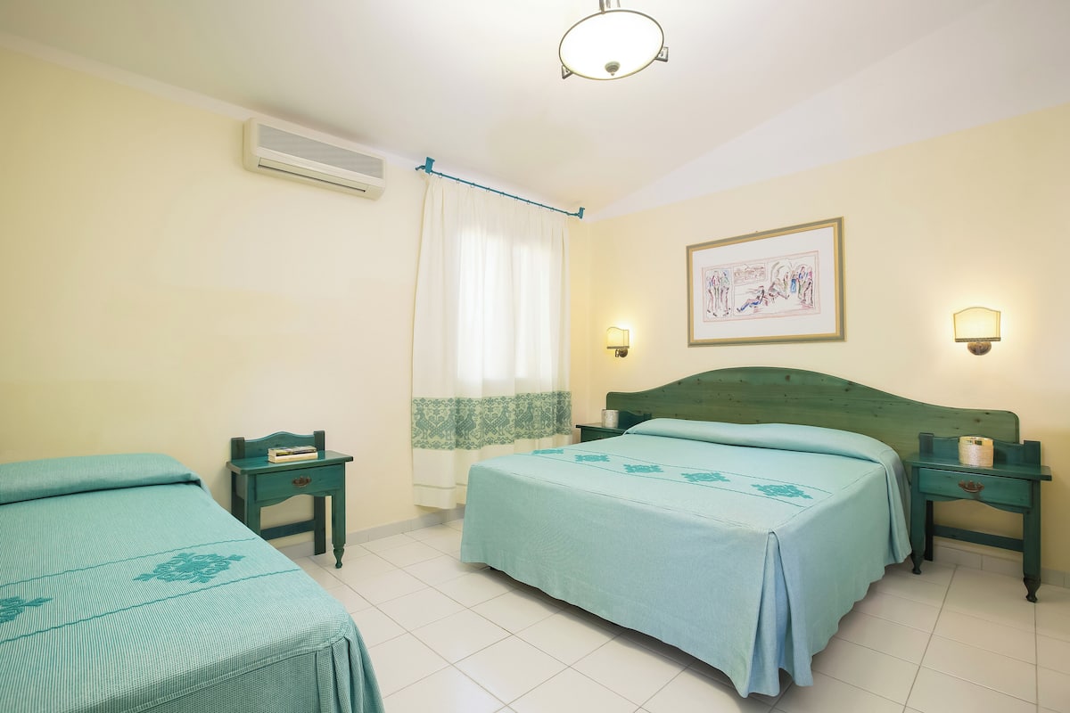 A hotel room with two beds, a nightstand, a lamp, an air conditioner, a curtain, and a framed picture on the wall.