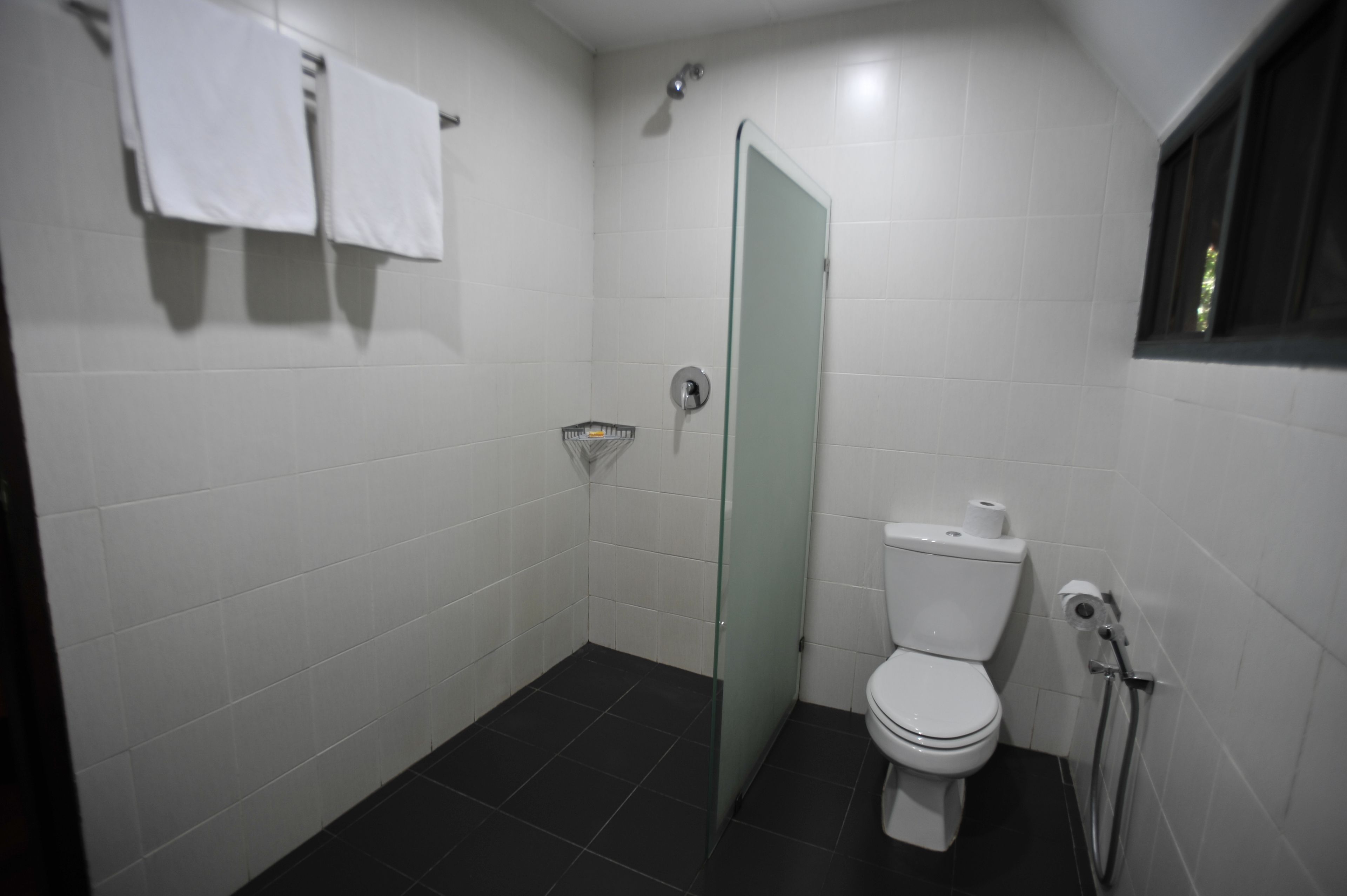 Un baño pequeño y limpio con azulejos blancos, cabina de ducha de vidrio, inodoro y ventana.