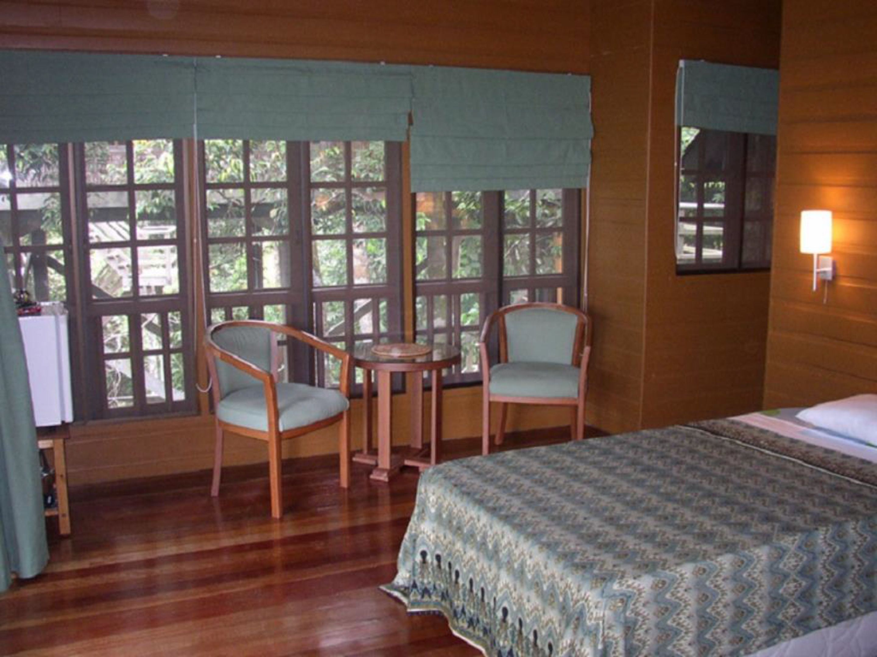 A room with a bed, two chairs, a small table, and large windows with a view of trees.