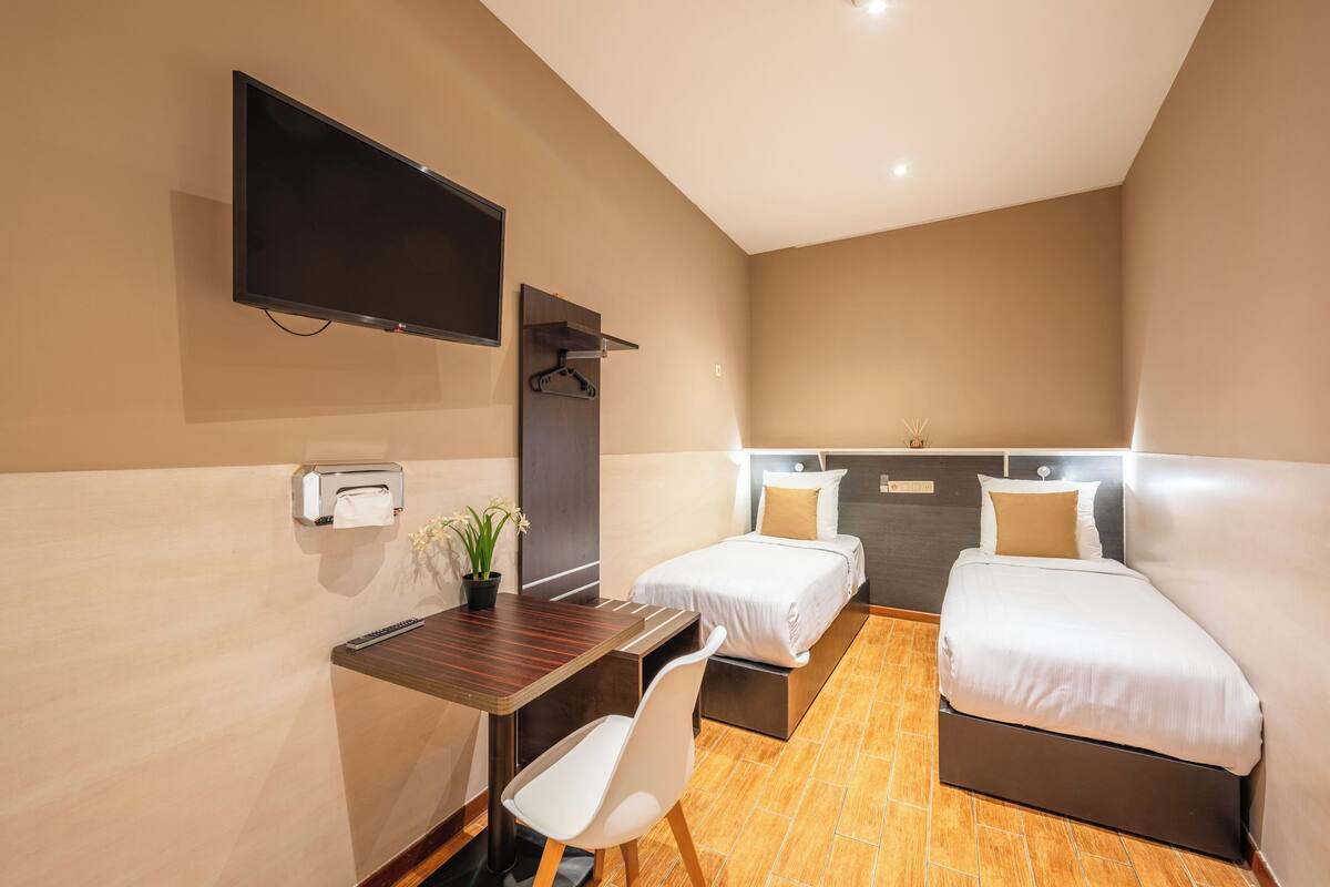 A hotel room with a bed, a desk with a chair, a TV mounted on the wall, and a small table with a plant.