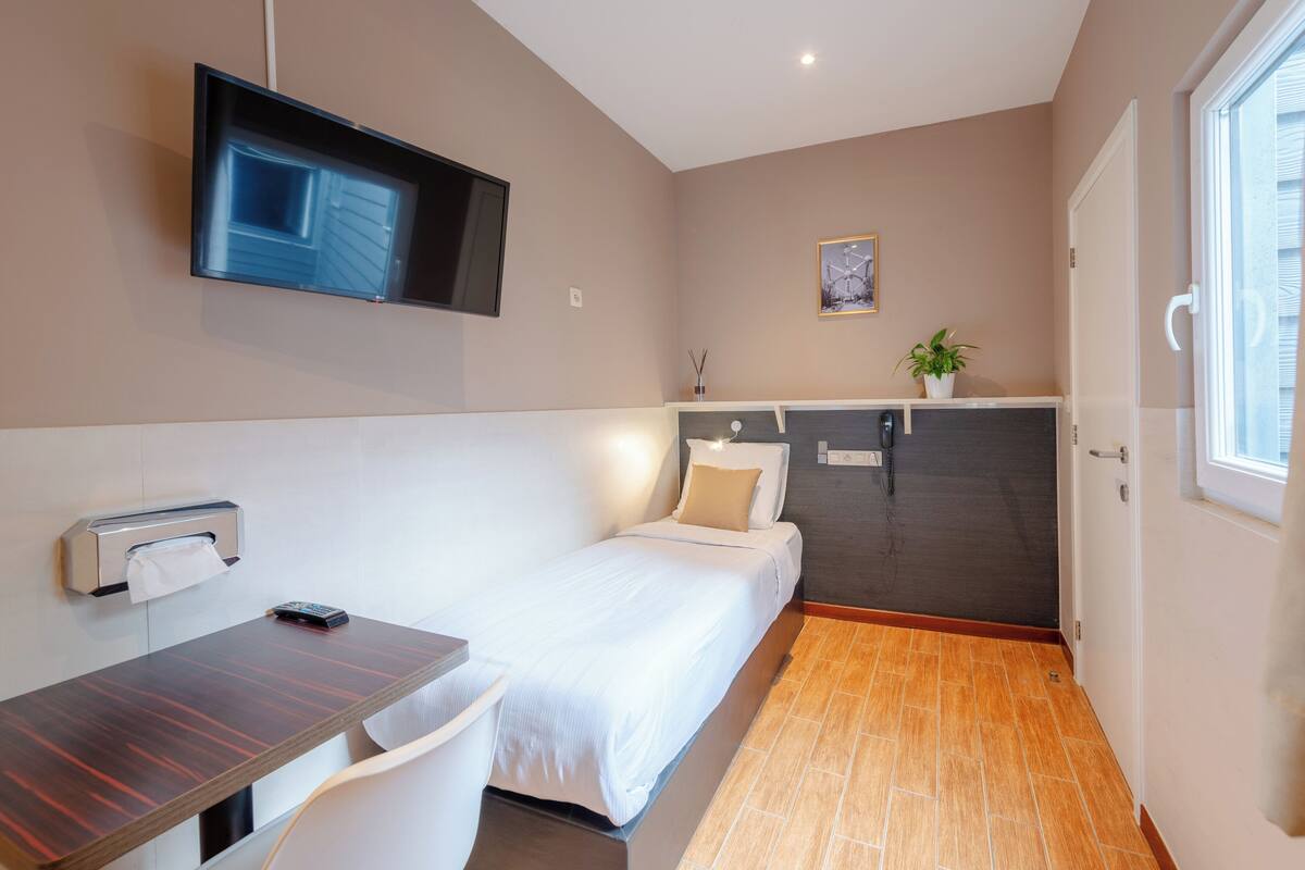 A compact hotel room with a bed, a desk, a TV mounted on the wall, a small window, and a door leading to a bathroom.