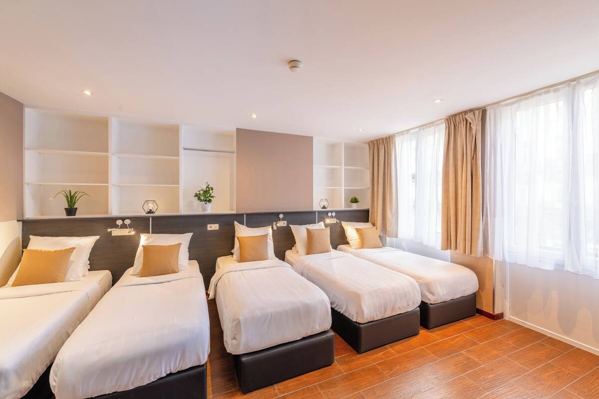 A hotel room with three beds, a large window with curtains, a wall-mounted shelf with plants, and a ceiling light.