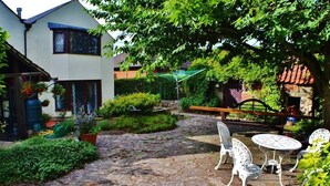 A stone-paved courtyard with white metal chairs and a table, surrounded by greenery and a house with a blue roof. - Church Villa B&B (Chesterfield)