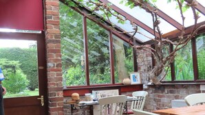 A cozy room with a brick wall, a wooden table, and white chairs. A large window offers a view of greenery outside. - Church Villa B&B (Chesterfield)