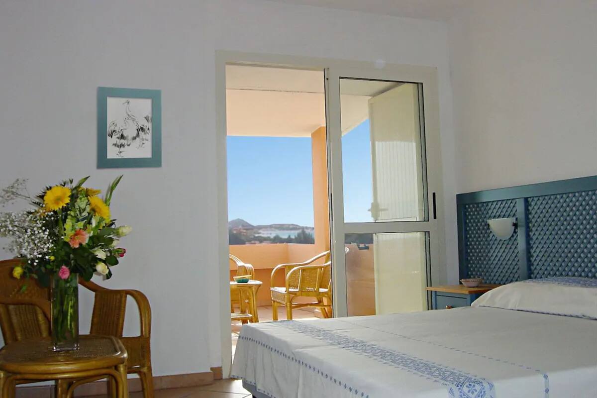 A hotel room with a bed, a chair, a table, a vase of flowers, and a balcony with a view.