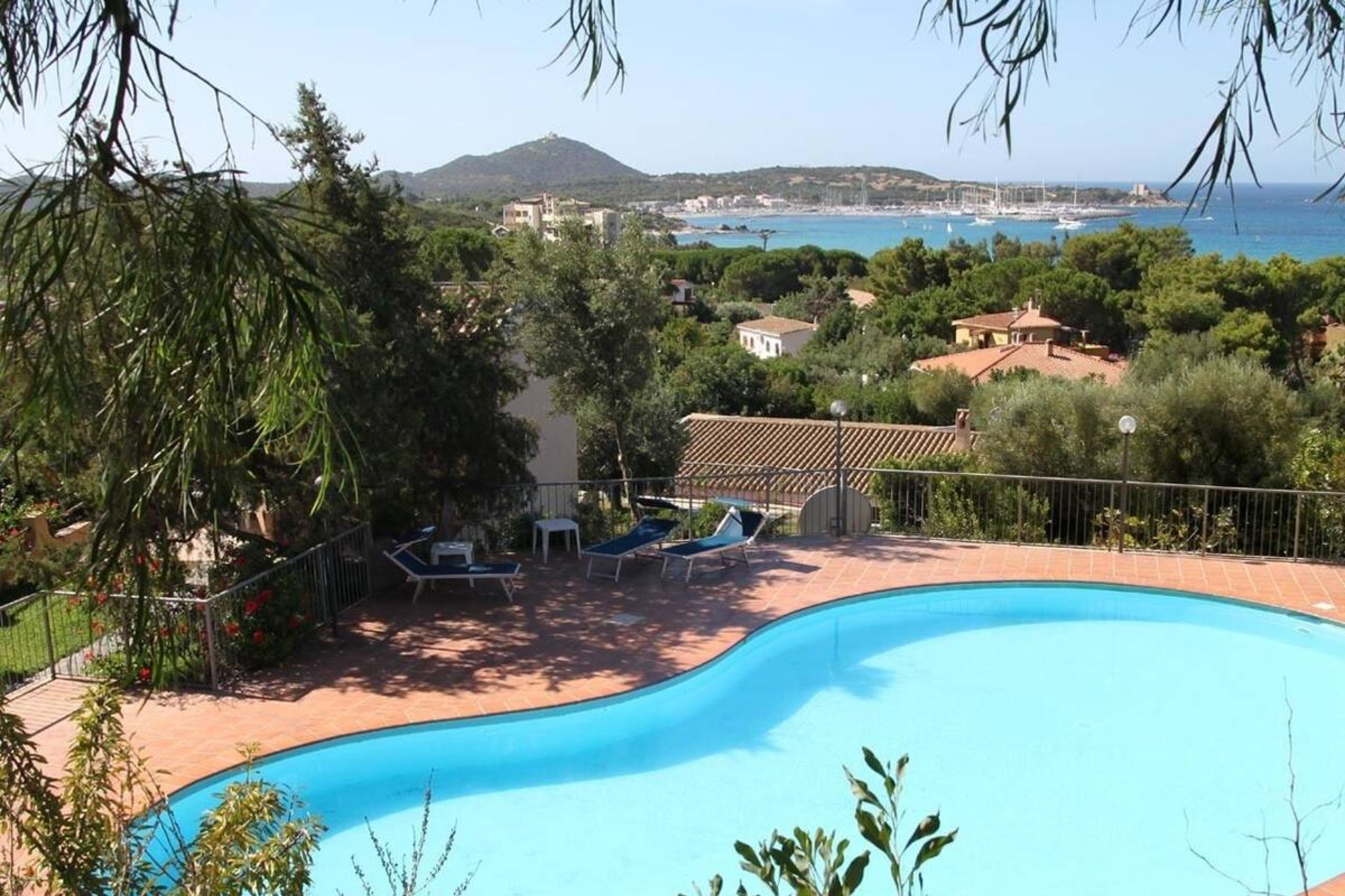 A swimming pool with lounge chairs, surrounded by greenery and overlooking a coastal area with buildings and a mountain in the distance.
