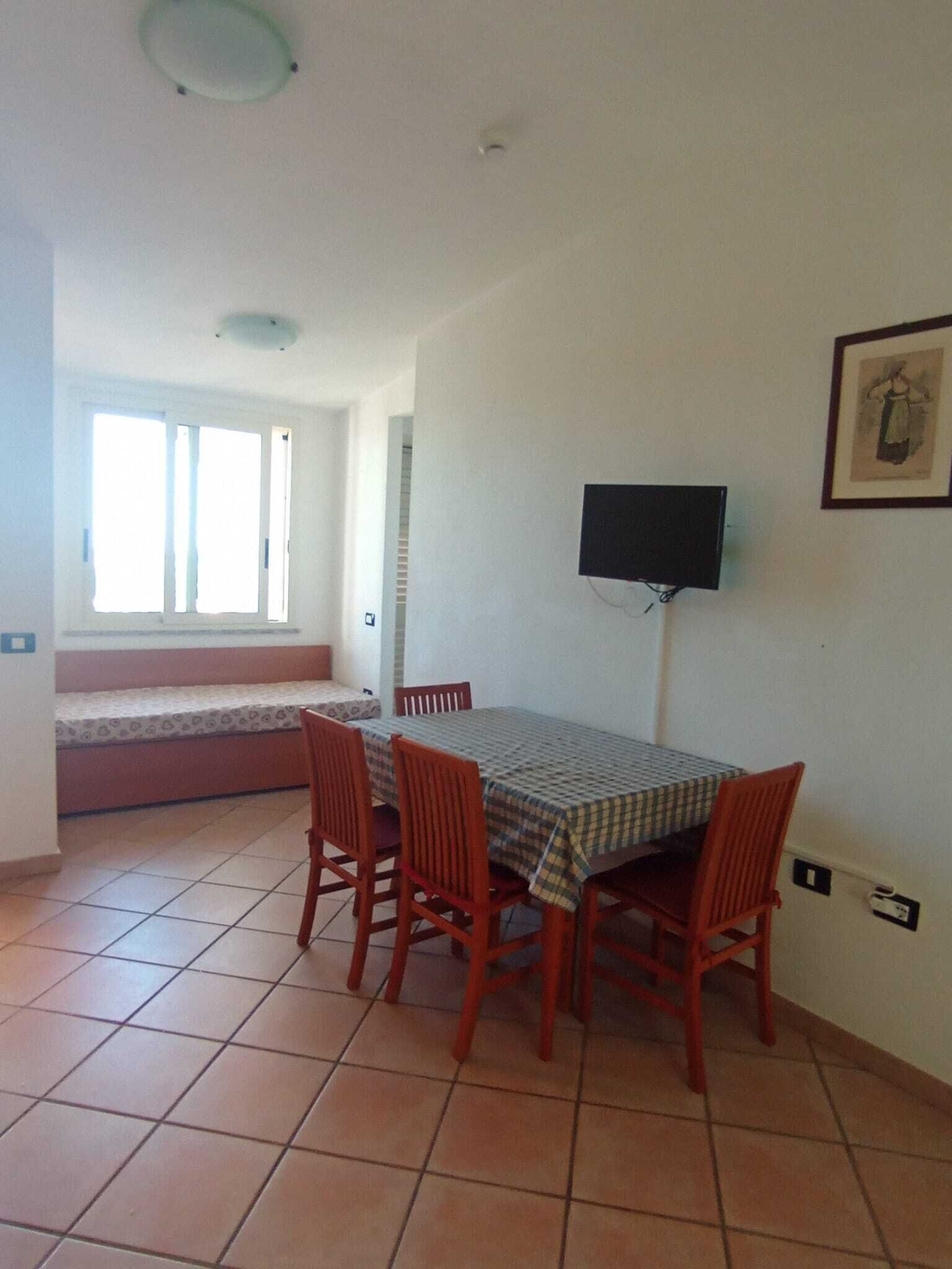 A dining area with a table and chairs, a television mounted on the wall, and a window with blinds.