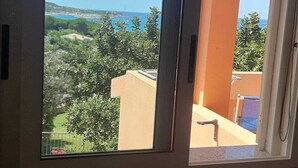 A room with a large window offering a view of a green landscape and a body of water. - Residence la Chimera (Villasimius)