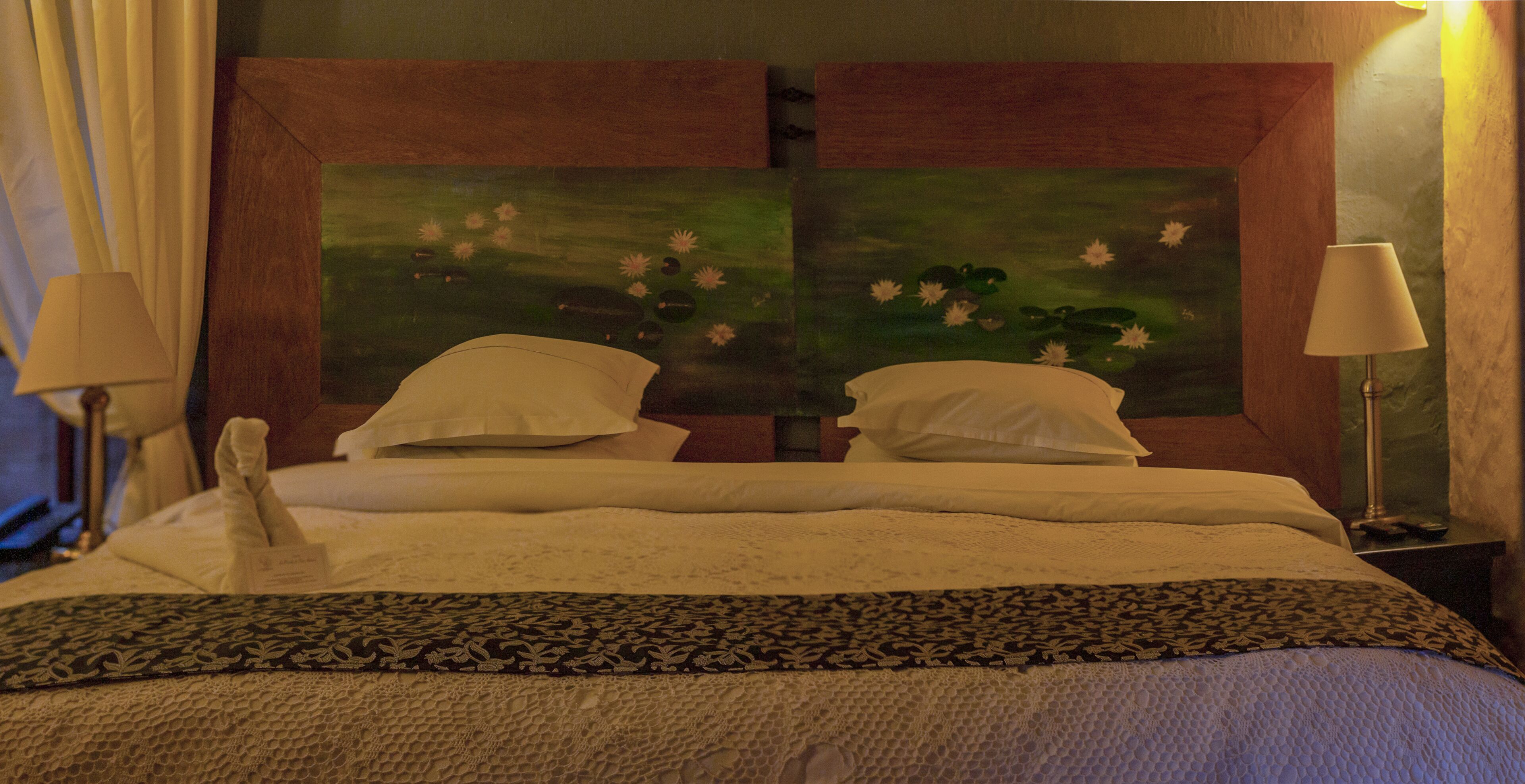 Habitación de hotel con una cama grande, dos almohadas, una mesita de noche con lámpara y un cuadro en la cabecera.