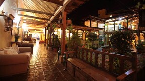 A cozy, rustic interior with wooden beams and a stone floor. There are cushioned seating areas and potted plants.